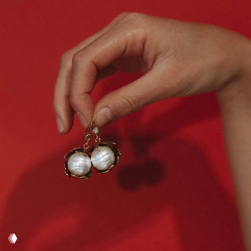 Рука держит серьги с натуральными барочными жемчужинами на красном фоне, показ формы жемчужин, блеска и декоративной оправы изделия.