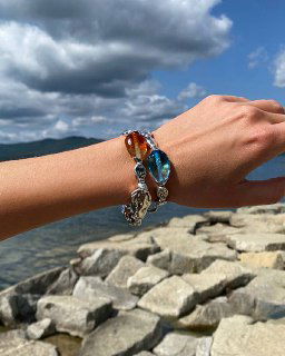 Запястье с двумя браслетами Ciclon LIQUID на фоне каменистого побережья: металлическая цепь и янтарные стеклянные элементы гармонируют с морской темой.