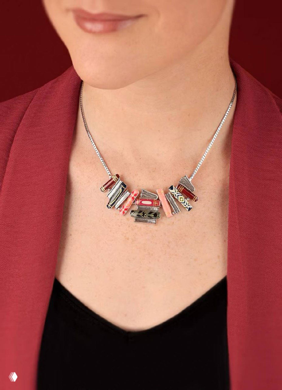 Колье TARATATA ROMANS на шее модели: разноцветные миниатюрные «книжки» на тонкой серебристой цепочке, показано в образе с бордовым жакетом и нейтральным макияжем.