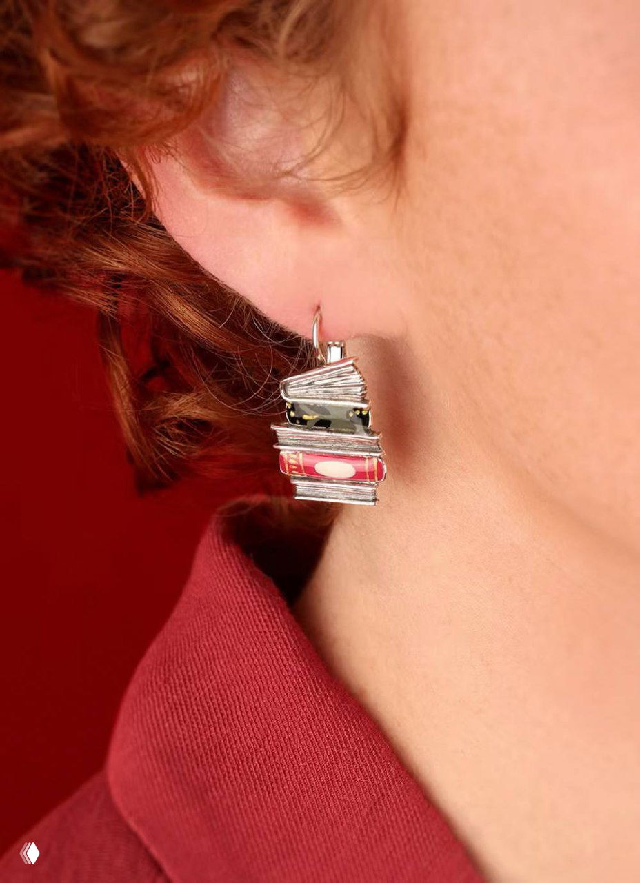 Серьга ROMANS TARATATA в ушной мочке модели, боковой ракурс: стилизованные сложенные книжные элементы, серебристая основа и разноцветная эмаль в деталях.