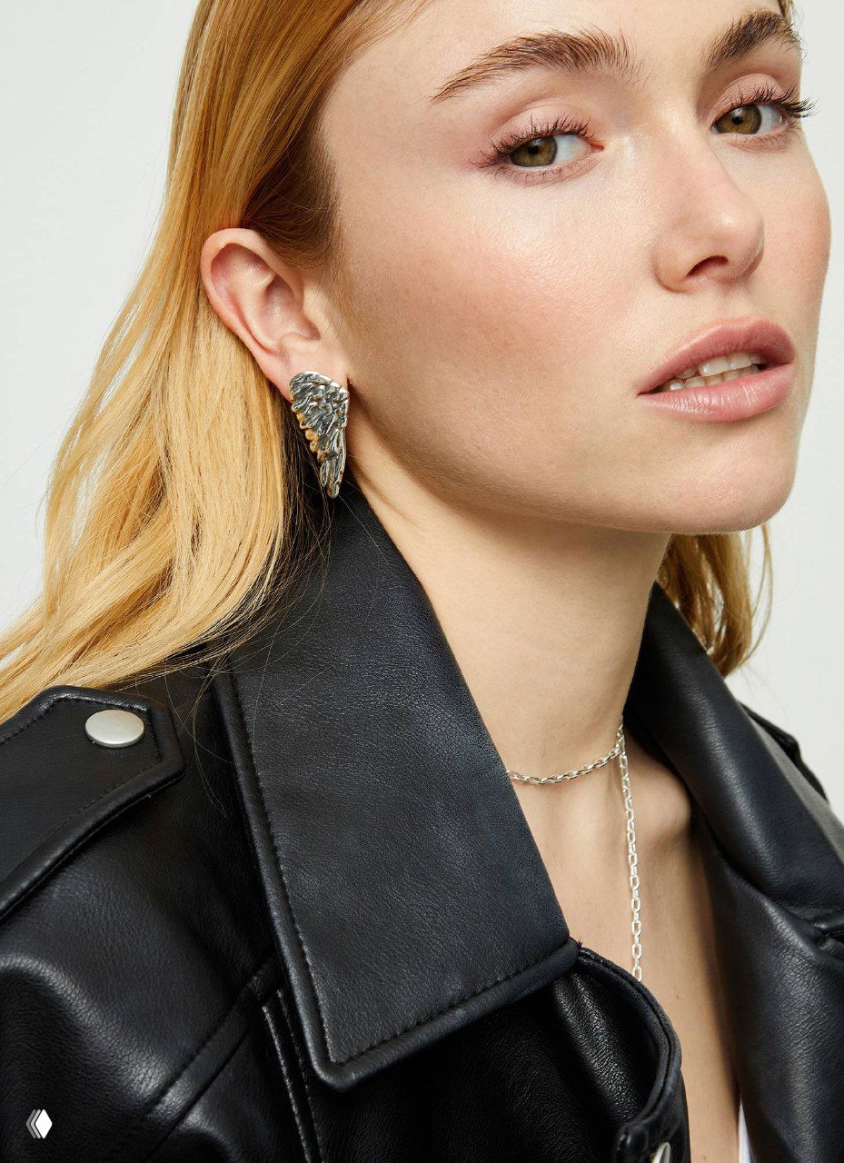 Крупный план модели в чёрной кожаной куртке: серебристая серьга в форме крыла и тонкая цепочка — каталоговый снимок новинки UNOde50.