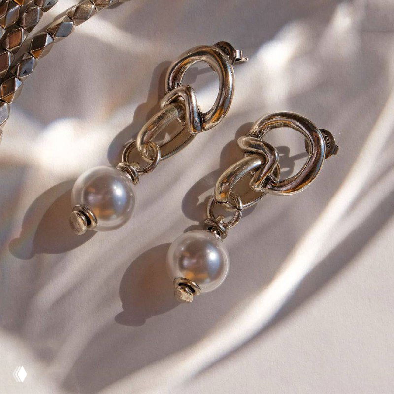 Крупный план серьг с подвесными жемчужинами UNOde50 на светлом фоне — детальное отображение отделки и отражений металла.