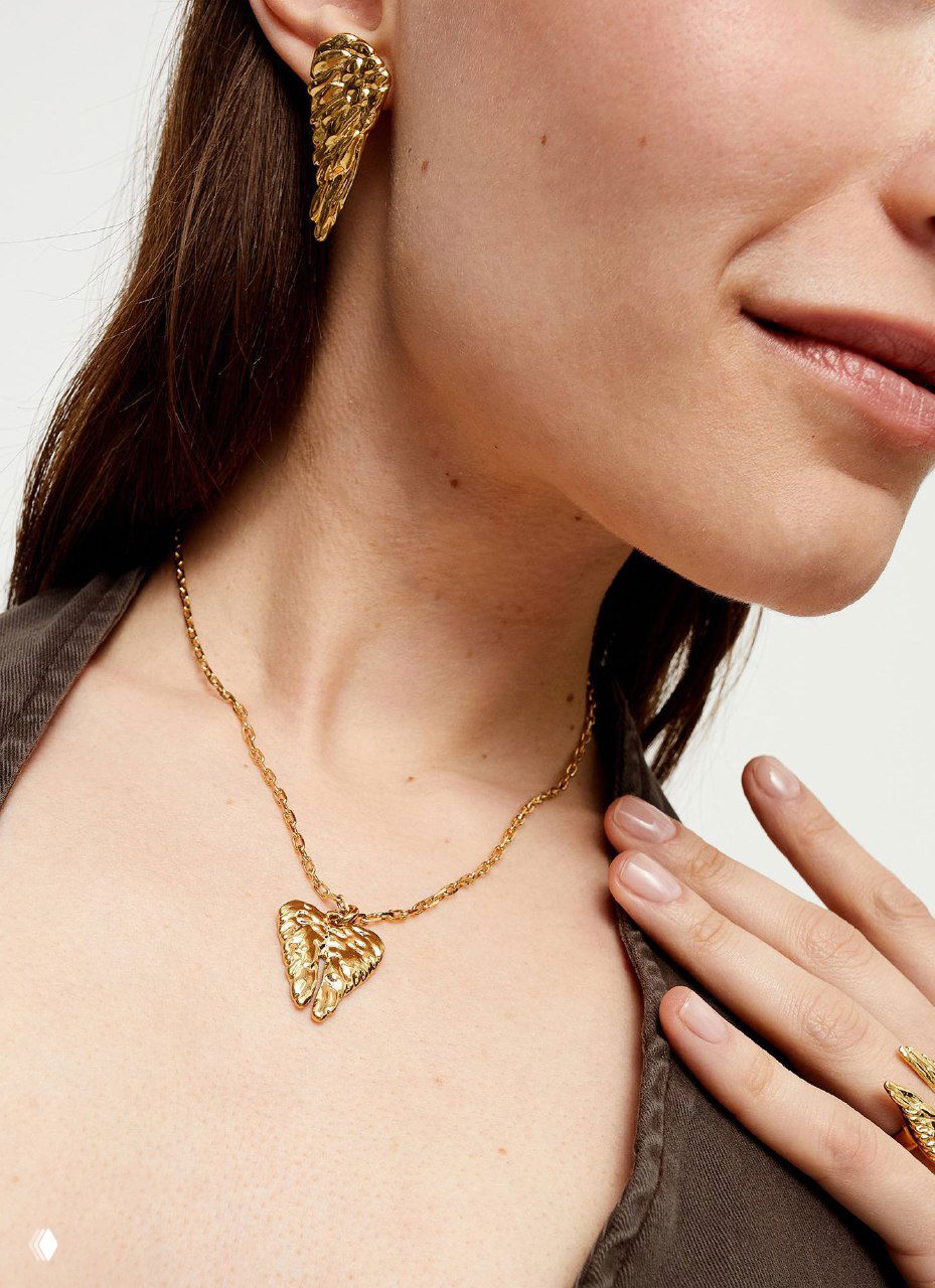 Крупный план украшений: золотые серьги и кулон в форме крыла на шее модели — стилизованная съёмка коллекции SER INVENCIBLE.