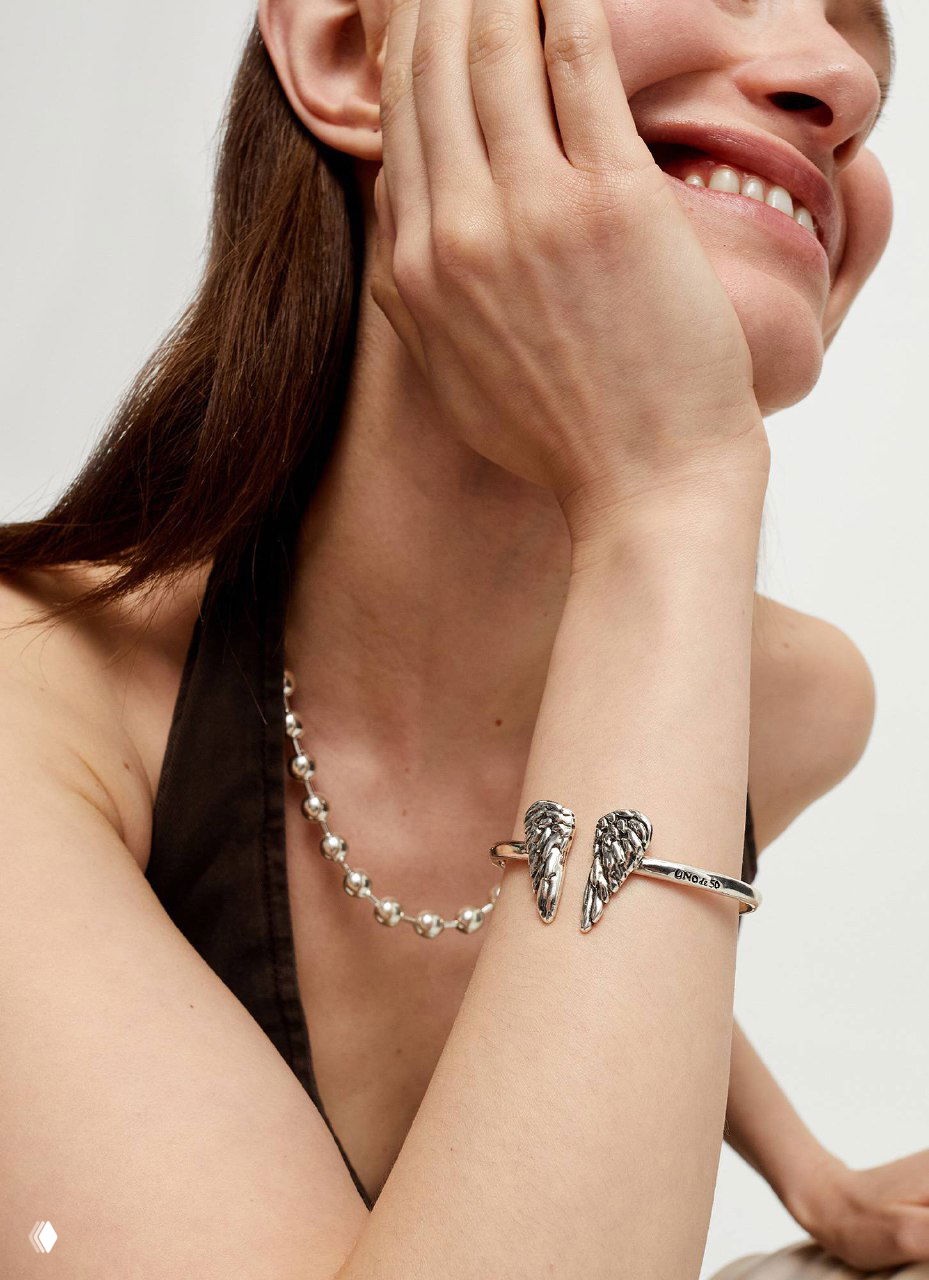 Модель демонстрирует браслет с декоративными крыльями и тонкую жемчужную цепочку на шее — стилизованный снимок коллекции UNOde50.