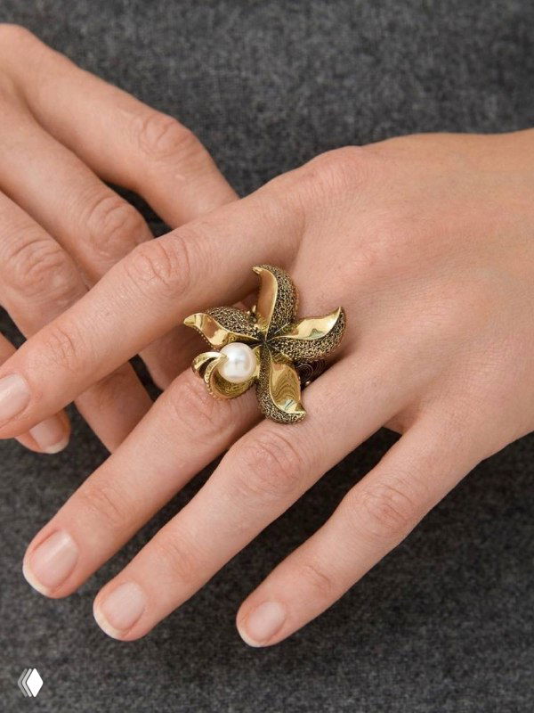 Крупный план пальцев с кольцом-цветком Fiore di Firenze и жемчужиной, фактурная латунь и сияющий перламутр на нейтральном фоне.