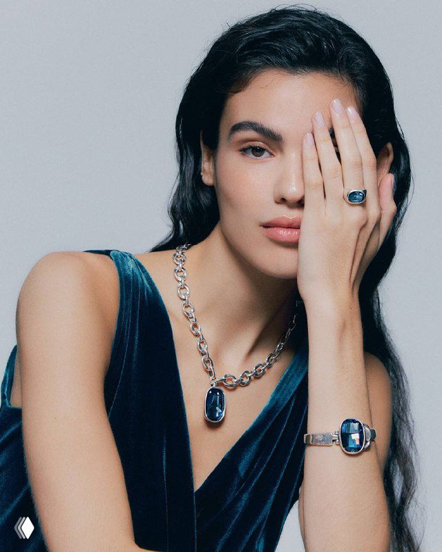 Женщина в синем платье демонстрирует колье и кольца UNOde50 с синими вставками — сочетание украшений в образе и стилистике бренда.
