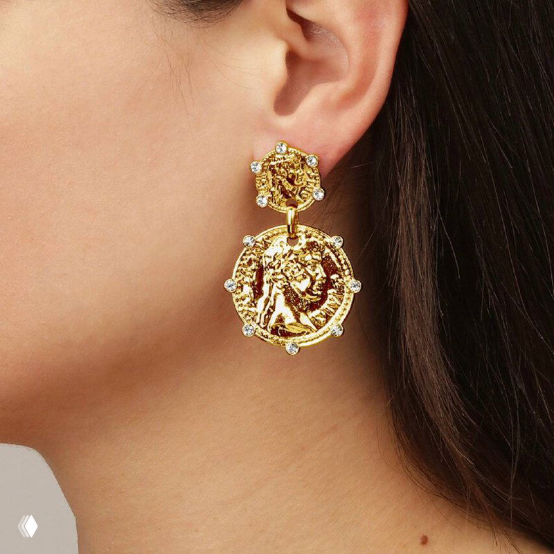 Крупный план серьги Dyrberg Kern в золотистом тоне с рельефной медальонной деталью и мелкими инкрустациями; на ухе модели видно посадку украшения.