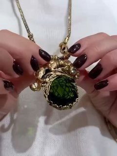 Крупный план: женские руки с тёмным маникюром держат золотую подвеску с крупным зелёным камнем и декоративными деталями коллекции «Сафари».