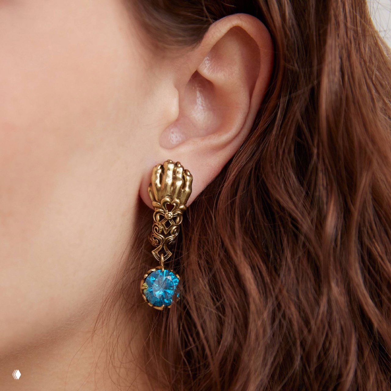 Крупный план серьги на ухе: декоративный элемент в форме лапы и подвес с ярко-голубым кристаллом на фоне тёмных волос модели.