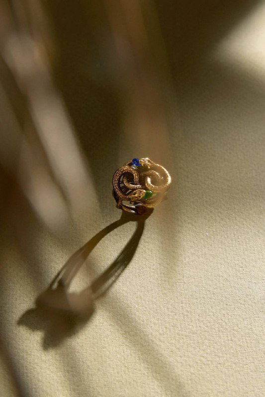 Крупный план маленького золотого кольца‑змеи со вставкой синего камня, тёплый свет создаёт выразительную тень от украшения