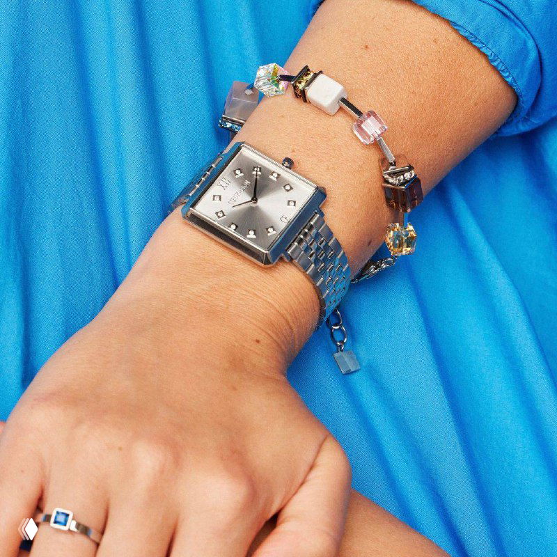 Крупный план женского запястья в голубом платье: серебристые часы Coeur de Lion на браслете рядом с декоративным браслетом и кольцом, стильная съемка.