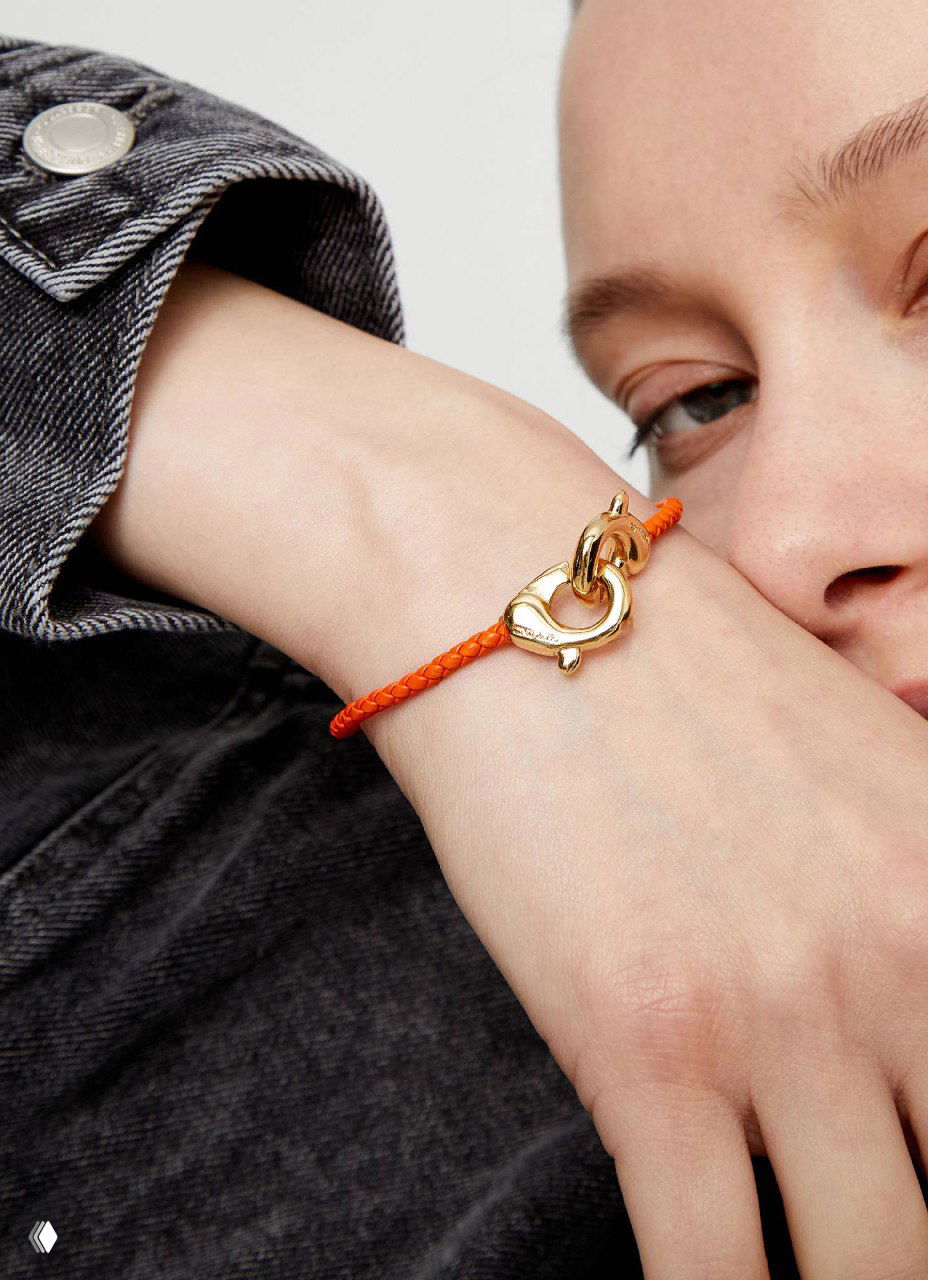 Крупный план женского запястья в оранжевом тонком кожаном браслете с позолоченным карабином — сочетание цвета и металлического акцента.
