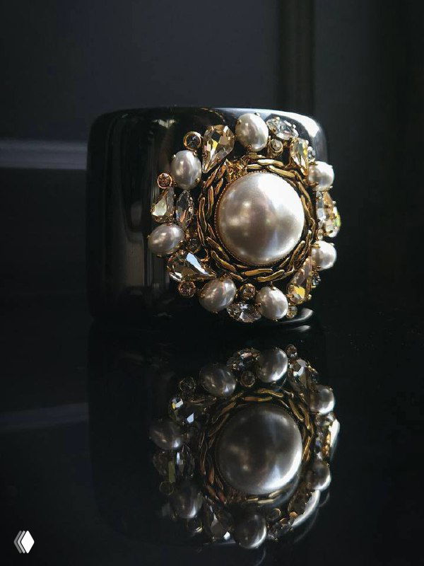 Чёрный широкий браслет с крупной центральной жемчужиной и мелкими декоративными элементами в золотой оправе, отражение на глянцевой поверхности.
