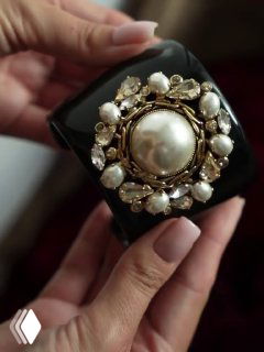 Детальный кадр чёрного браслета с жемчугом в руке; видны текстура материала, золочёная фурнитура и декоративные кристаллы вокруг центральной жемчужины.