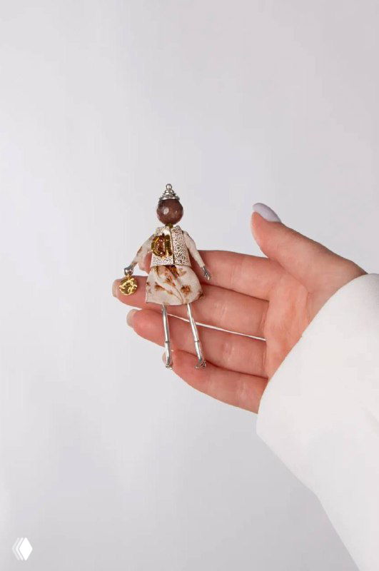 Брошь-кукла MIAMELIE в руке на белом фоне: видны тканевые детали, металлические элементы и декоративные вставки, аксессуар в натуральном виде