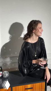 Сидящая женщина в вечернем платье с длинными серьгами и ожерельем, естественный свет создаёт тёплую атмосферу и тени на стене.