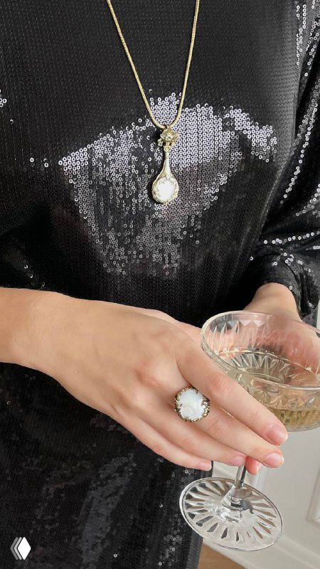 Макро-снимок ожерелья с крупной белой подвеской и рук с кольцами, модель держит бокал — подчёркнуты материалы и дизайн изделий.