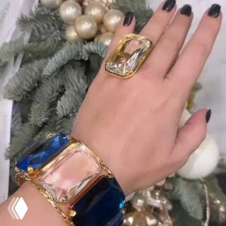 Крупные прямоугольные прозрачные и тёмно-синие камни в золотой оправе на руке на фоне праздничной ёлки — демонстрация коллекции украшений.