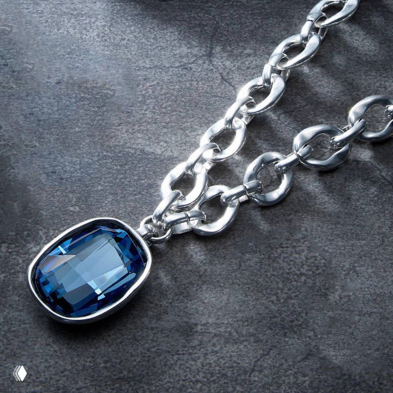 Крупный план серебряного кулона UNOde50 с крупным синим кристаллом на массивной цепочке, студийная съемка на темном фоне для каталога украшений