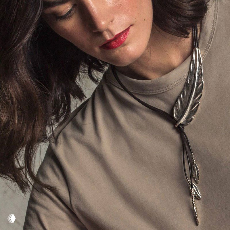 Модель в бежевой одежде с удлинённым подвесом в виде пера от UNOde50, фото образа для каталога с акцентом на стилизацию и детали украшения