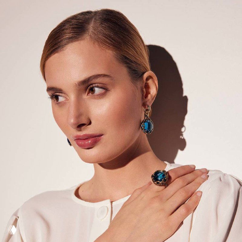 Модель в светлой блузе демонстрирует серьги и кольцо CATENE: сине-зеленое сияние камней и золотистая цепная оправа.