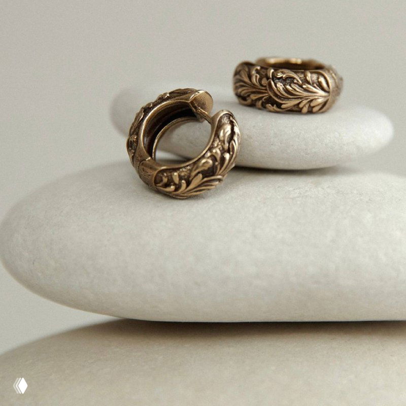 Пара округлых серьг с резным растительным орнаментом на светлом монохромном фоне, демонстрация формы и деталей металла в винтажном стиле.