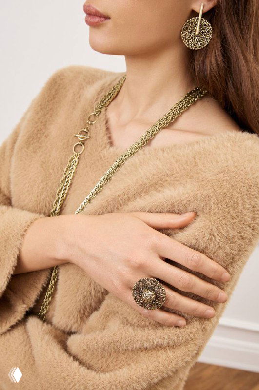 Модель в бежевом свитере демонстрирует металлические украшения: массивное колье, крупные серьги и кольцо, подчёркивающие блеск металла и текстуру деталей.