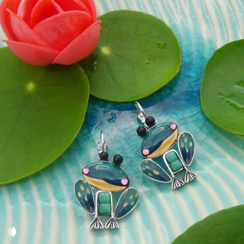 Яркие подвесные серьги‑лягушки TARATATA с зелёной и бирюзовой эмалью на фоновой композиции с листом и цветком, декоративный летний стиль.