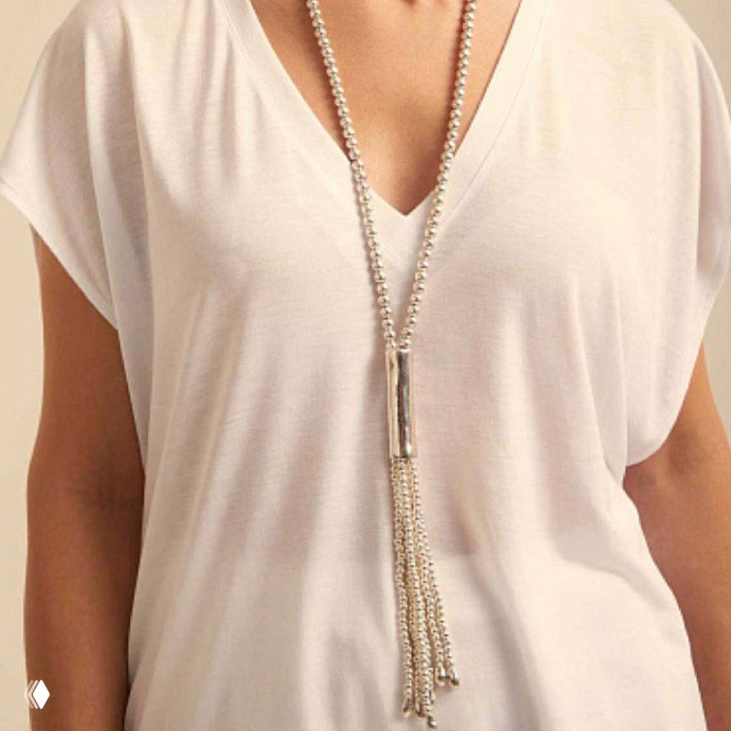 Длинное ожерелье UNOde50 с кисточкой и серебристыми элементами на фоне светлой блузы, стилизованная демонстрация образа.