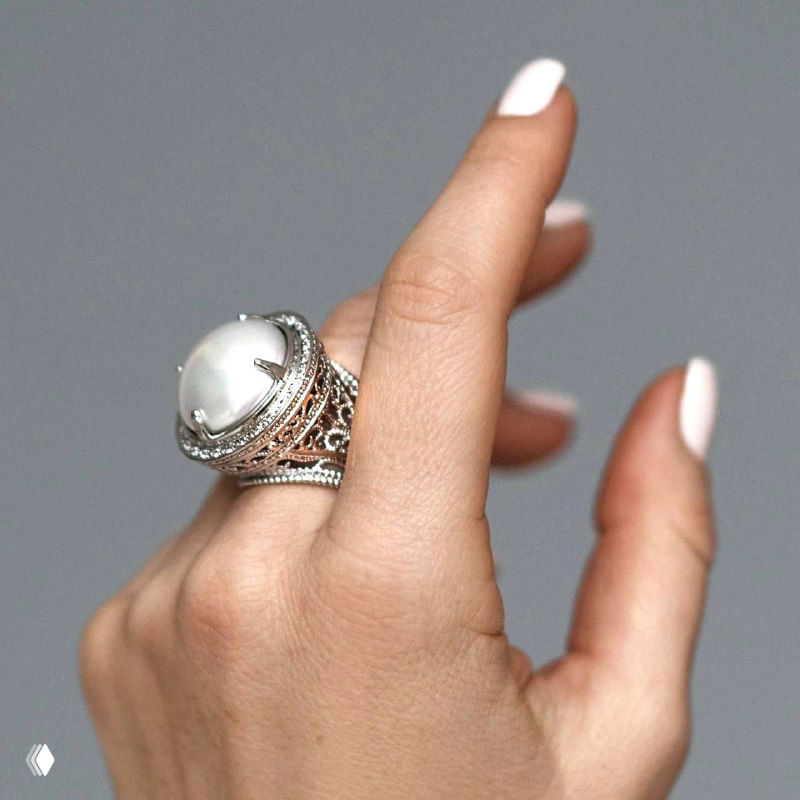 Крупный серебристый коктейльный перстень с овальным светлым камнем на согнутой руке с аккуратным белым маникюром, студийная съёмка.