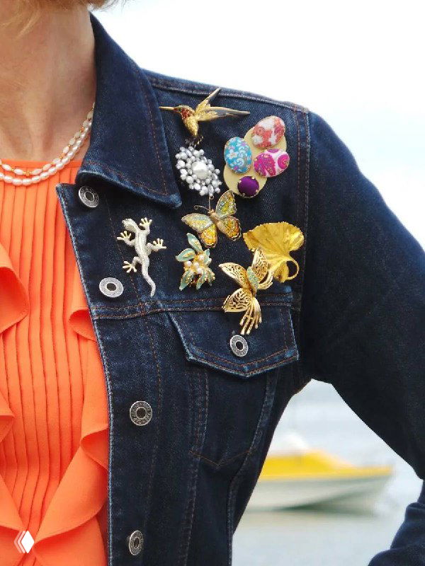 Детальный кадр джинсовой куртки с композицией разноцветных брошек на лацкане — цветочные мотивы, птицы и блестящие вставки в модном образе.