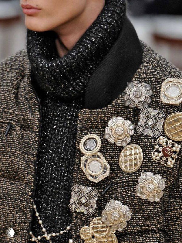 Крупный план твидового пальто с аккуратно сгруппированными декоративными брошками разных форм и металлов — текстура ткани и детали украшений видны крупно.
