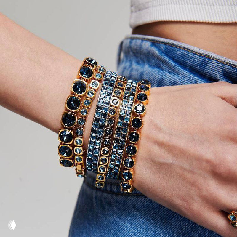 Несколько браслетов на запястье: комбинация тонких и массивных браслетов с тёмно‑синими кристаллами и золотистой фурнитурой на фоне джинсовой одежды.