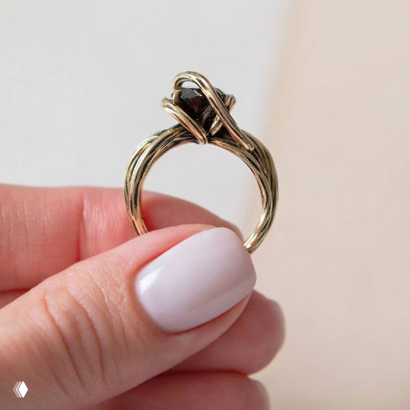 Крупный план: кольцо CORDA золотистого оттенка в пальцах, перекрученные ленты и винтажный дизайн на светлом нейтральном фоне