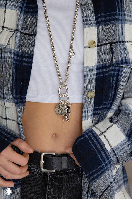 Модель в клетчатой рубашке и белом топе демонстрирует длинную подвеску Ciclon на цепочке с декоративными брелоками в модном образе.