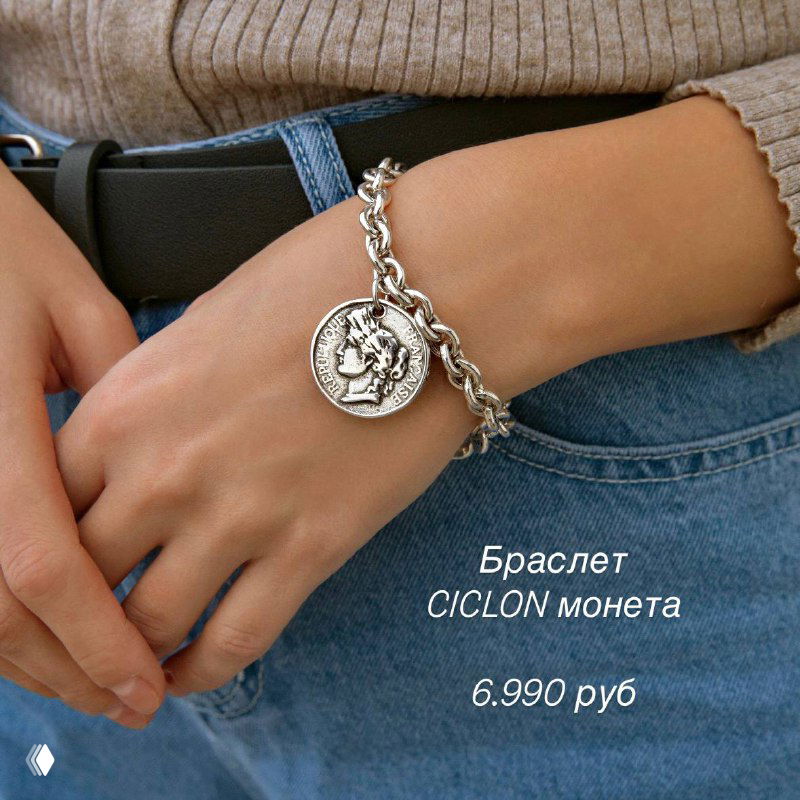 Крупный план запястья в джинсах и ремне с массивным серебристым браслетом Ciclon с подвеской-монетой, показан стиль и цена товара.