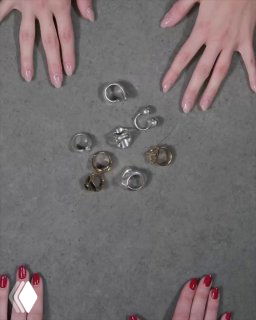 Набор серебристых и золотистых колец UNOde50, разложенных на сером фоне в центре, рядом видны руки с маникюром