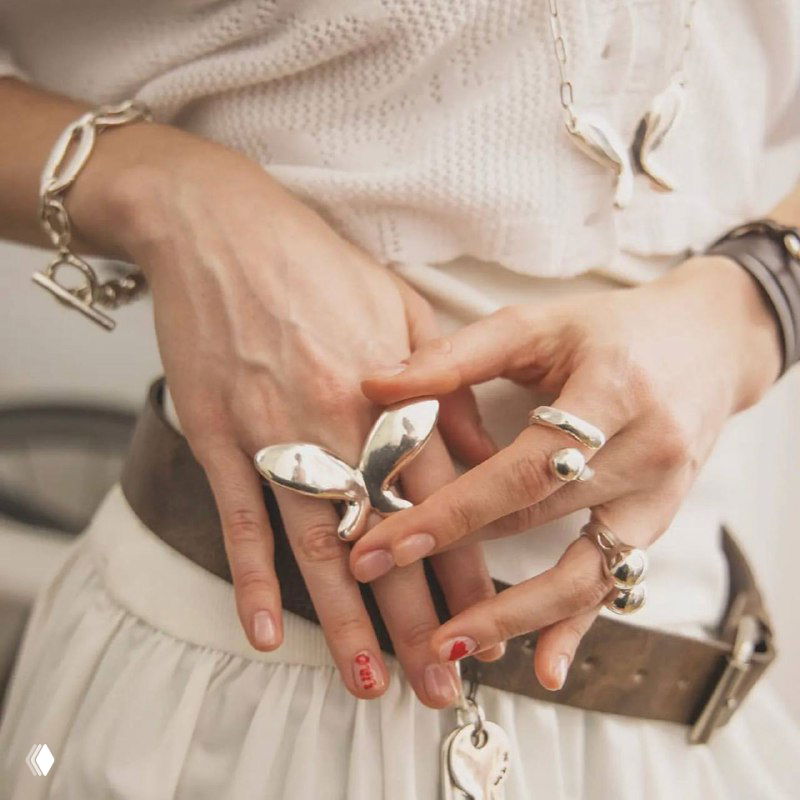 Женские руки в светлой одежде с несколькими крупными фигурными кольцами UNOde50 на пальцах и браслетом — образ в студийном стиле