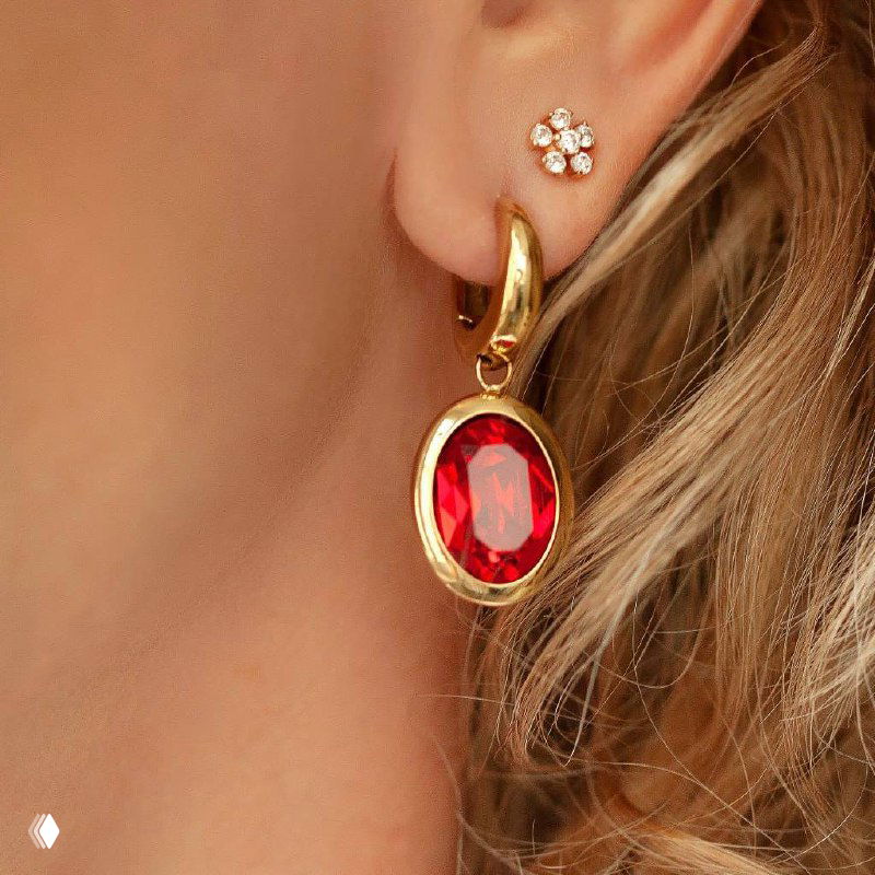 Крупный план позолоченной серьги QUDO с овальным красным кристаллом на фоне светлых волос, акцент на форму и блеск