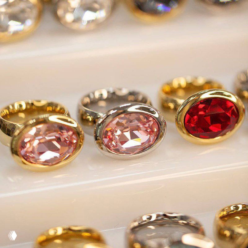 Ряд позолоченных колец QUDO с крупными круглыми кристаллами розового и красного оттенка на выложенной подставке