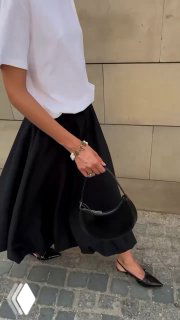 Уличный кадр: модель в белой футболке и чёрной юбке, держит сумку; на запястье видно браслет с жемчужными деталями — повседневный стиль с украшениями.