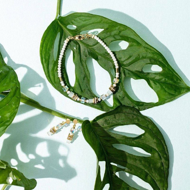 Кремовый и золотой комплект Coeur de Lion — браслет и серьги, аккуратно уложенные на крупных зелёных листьях в студийной выкладке для летнего образа.