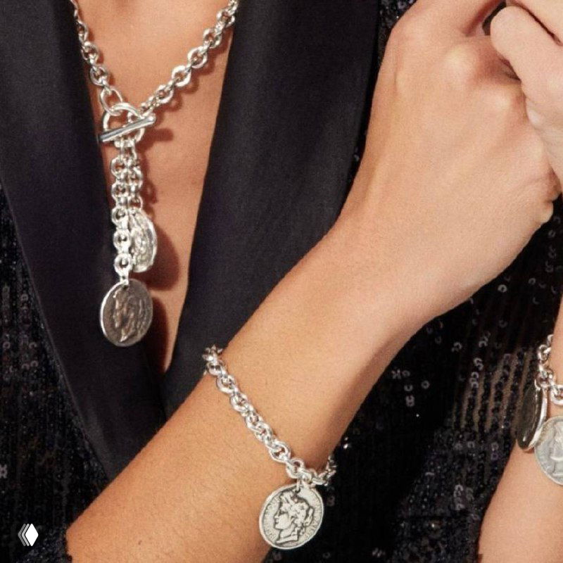 Крупный план браслета Ciclon с подвеской-«монетой» на запястье модели, серебристая цепочка в сочетании с чёрным пиджаком, стильный кадр.