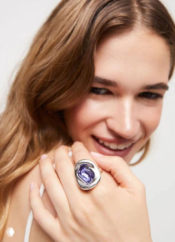 Модель улыбается и показывает крупное кольцо UNOde50 с фиолетовым кристаллом из коллекции Ser Valiente на руке, студийная съемка.