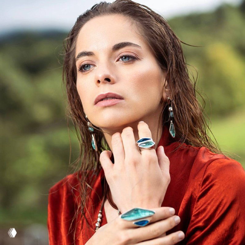 Модель в красном бархатном платье демонстрирует серьги и кольцо с бирюзовыми элементами муранского стекла на природном фоне.