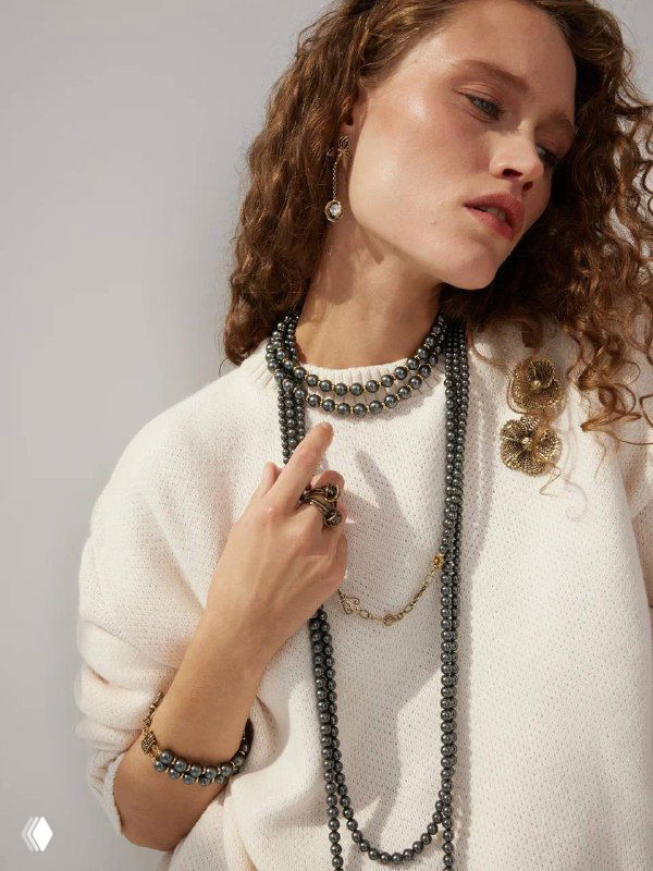 Модель в белом вязаном свитере демонстрирует несколько длинных тёмно-серых жемчужных и золотых цепочек, браслет и декоративную брошь в образе.