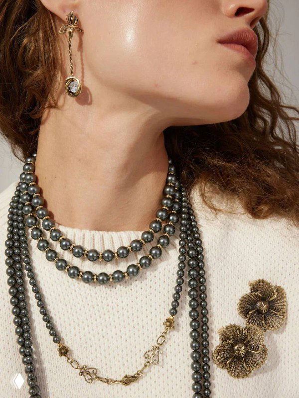 Крупный план шеи модели с многослойными тёмно-серыми жемчужными ожерельями, золотой цепочкой и изящной серьгой на фоне белого свитера.