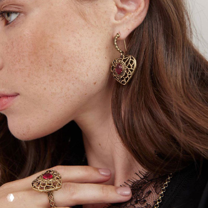 Крупный план профиля модели в серьге‑сердечке с красным камнем и детальной ажурной работой металла, сочетание с кольцом на пальце.