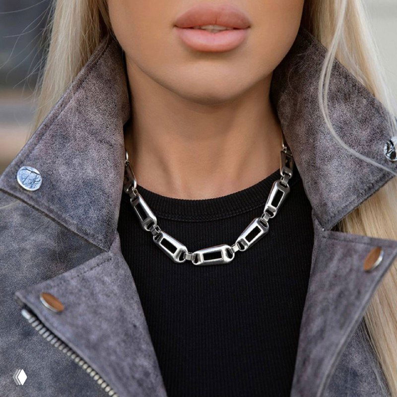 Женская модель в светлой куртке и чёрном верхе носит массивное серебристое колье-цепь на шее — уличный образ с акцентным украшением