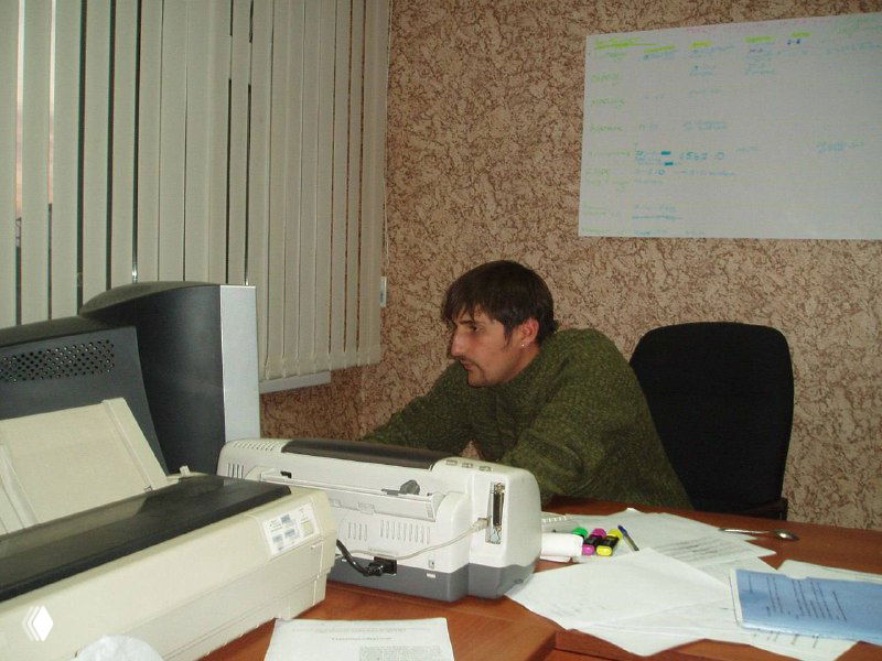 Фотография: молодой мужчина за офисным столом с монитором и принтером, рабочие бумаги на столе — снимок из эпохи 2000-х.